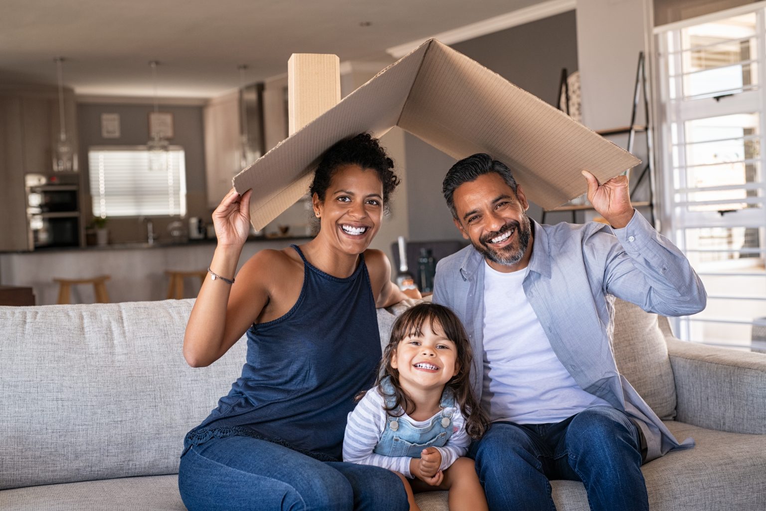 Portrait of smiling family sitting on couch holding cardboard roof and looking at camera. African and indian parents with funny daughter holding cardboard roof over heads while sitting on sofa in their new home. Happy mother and father with daughter in new house at moving day with copy space.